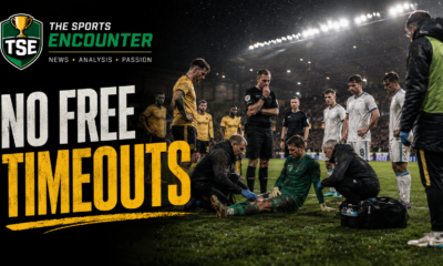 Goalkeeper receives treatment during a tense soccer stoppage as players, match officials, and bench staff wait under stadium floodlights, with The Sports Encounter branding and the headline “No Free Timeouts.”